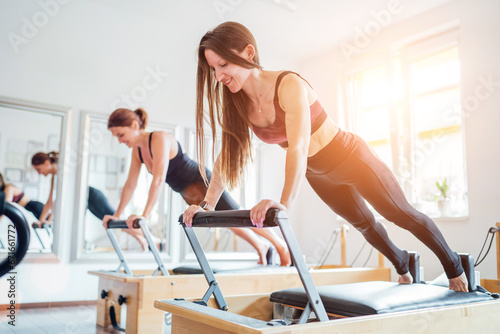 Two young females doing extended plank static strengthening core muscles exercise using pilates reformer machine in sport athletic gym hall. Active people training, yoga classes concept.