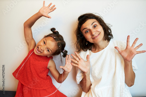 Babysitter and little girl in red dress having fun playing together at home against white wall, grimacing to frighten you, child making scary face, woman looking at camera with big eyes and smile