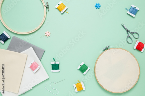 Embroidery set fot stitching. Beige cotton cloth in embroidery hoop on green background with fabric, colorful threads, scissors and needls. Indoor hobby concept with copy space, top view