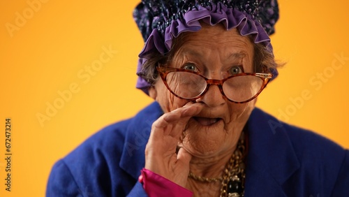 Closeup funny portrait of smiling happy crazy toothless grandmother with wrinkled skin puts hand to mouth to tell a secret isolated on yellow background studio