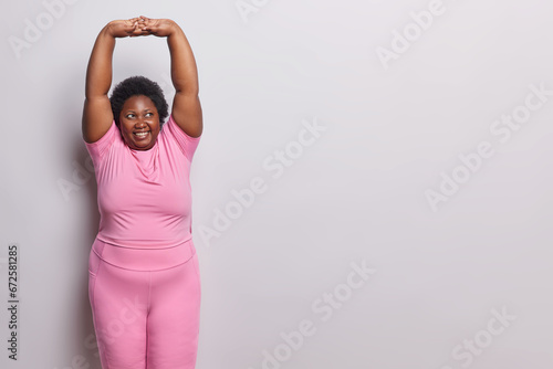 Sporty lifestyle. Indoor shot of young happy smiling plump darkskinned woman standing on left isolated on white background in pink tracksuit doing stretching exercise with blank space for promotion