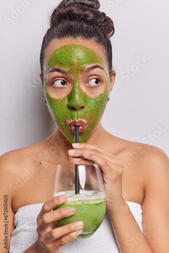 Vertical image of Latin woman applies green face mask while savoring smoothie radiating sense of wellnes focused aside has widely opened eyes wrapped in bath towel isolated over white background
