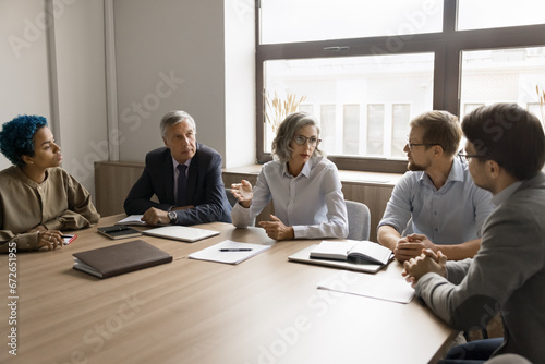 Serious mature business leader woman talking to diverse colleagues at office table, explaining tasks to employees. Multiethnic team meeting for conversation, collaboration, teamwork