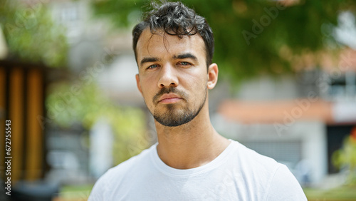 Young hispanic man standing with serious expression at park
