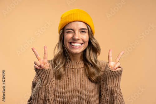 Cute smiling beautiful woman winking wearing stylish yellow hat showing victory sign isolated on beige background. Portrait of happy modern hipster female looking at camera. Positive lifestyle concept