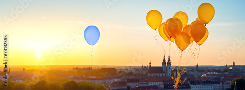 Standing out from the crowd concept. Blue balloon floating over a city separately from golden tied balloons during sunset
