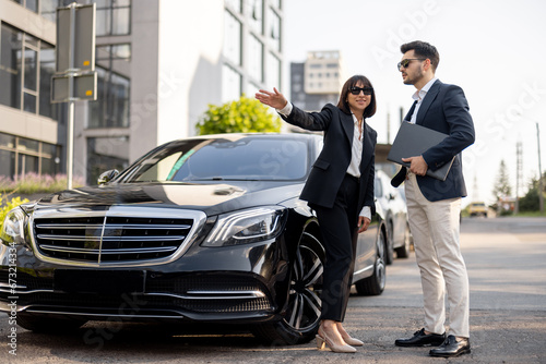 Business partners have a conversation while standing together near luxury car outdoors. Female driver showing way for a businessman arriving to destination