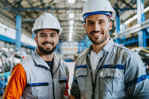 Zwei junge Männer Facharbeiter/Fachkräfte in Arbeitskleidung und Helmen lächelnd in einer Industriehalle