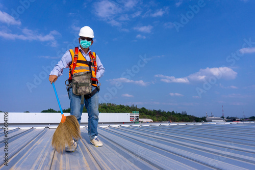 Worker wearing full safety body harness holding a broom in hand working on roof top for cleaning metal roof sheet