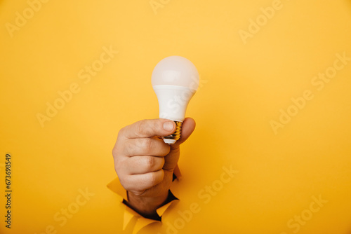 Hand holding an incandescent led light bulb from a torn hole in yellow paper