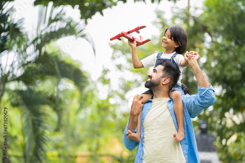 Happy smiling indian girl kid playing with airplane toy by sitting on father shoulder at park - concept of freedom, family support and togetherness