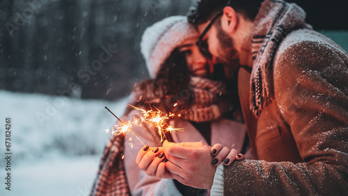 Outdoor waist up portrait of young beautiful happy smiling couple posing on street. Models hugging, looking at each other, holding sparkles, wearing stylish clothes. Snowfall. Copy, empty space