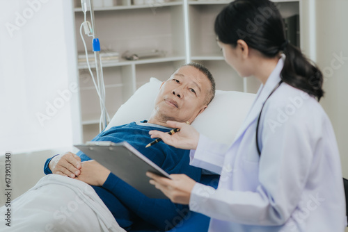 Beautiful Asian female doctor in white top talking with senior patient at bedside in hospital or clinic ward