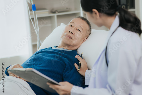 Beautiful Asian female doctor in white top talking with senior patient at bedside in hospital or clinic ward