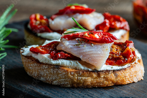 Two bread slices with ricotta cheese, pieces of herring and sun dried red tomatoes on wooden board background. Delicious breakfast or wine snack. Closeup