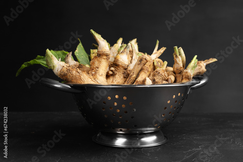 Fresh horseradish roots in colander on black textured table, closeup