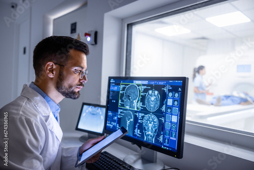 Doctor examining X-ray images on display in MRI control room while in background nurse preparing the patient for examination test.