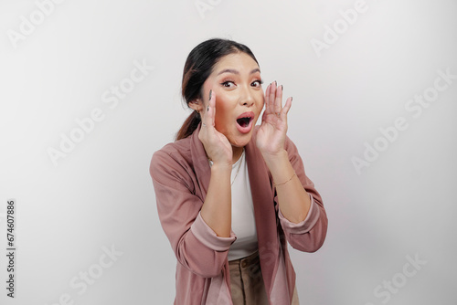 A young beautiful Asian woman employee wearing cardigan is shouting and screaming loud with a hand on her mouth, isolated by white background.