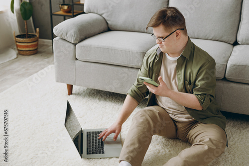 Young man with down syndrome wears casual clothes use laptop pc computer mobile cell phone sit grey sofa couch stay at home flat rest spend free time in living room. Genetic disease world day concept.