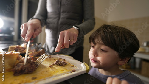 Little boy grabbing food by kitchen counter while mother prepare meal for family supper at night. Child observing mom cook food while grabs piece of meat