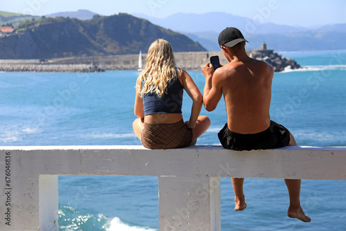jovenes pareja sentados mirando al mar con móvil desde un pretil zumaia verano getaria país vasco 4M0A2550-as23