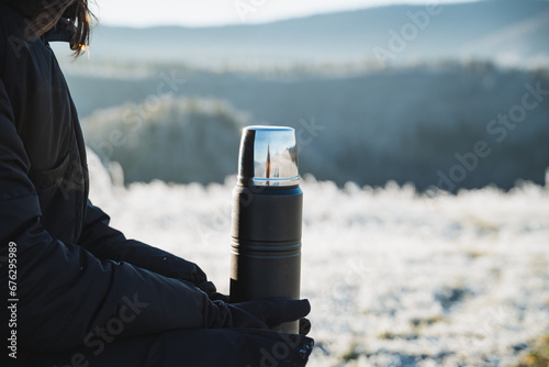 Black metal thermos on the background of winter forest, beautiful landscape, object in human hands, thermo flask for hot drinks.