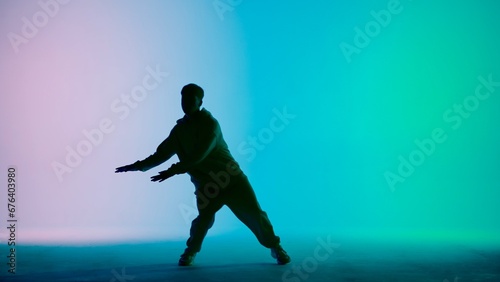 In the frame on a tricolor background, gradient in silhouette stands a young man. Demonstrates dance moves, stretching his arms forward. He is plastic, rhythmic. He is dressed in street style clothes