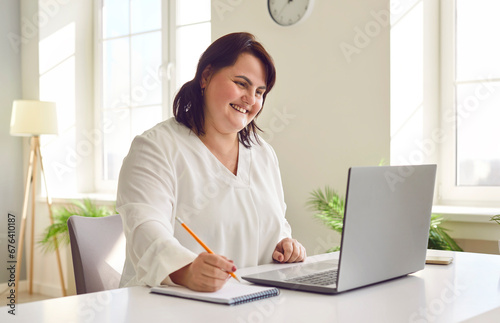 Smiling overweight young woman using laptop in office. Happy plus size woman reviewing financial documents and making notes in copybook. Financial manager, bookkeeper, accountant