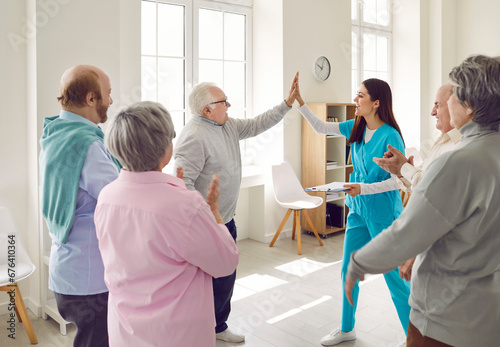 Senior man doing high five with a young friendly nurse in a group therapy for motivation. Elderly men and women standing in a circle in nursing home. Mental health for senior people.