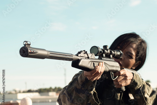 A female sniper with dark skin in camouflage military clothing holds a gun, takes aim, selective focus, optical sight close-up. Concept of military operations with the participation of female soldiers