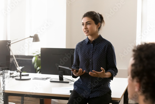 Serious pretty young Indian businesswoman leader talking to team on corporate meeting, speaking before employees, holding tablet, telling sales report result. Female business leader portrait