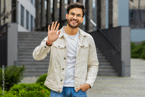 Hello. Indian man smiling friendly at camera, waving hands gesturing hi, greeting or goodbye, welcoming with hospitable expression outdoors. Happy Arabian Hindu guy standing in urban city town street