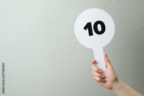 Woman holding auction paddle with number 10 on light grey background, closeup. Space for text