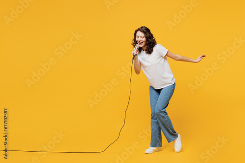 Full body singer happy fun young woman wear white blank t-shirt casual clothes sing song in microphone at karaoke club isolated on plain yellow orange background studio portrait. Lifestyle concept.