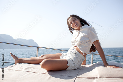 A beautiful young girl with a slender figure travels on a boat during a summer vacation in the open sea or ocean against the background of the island. A model is wearing a shirt and shorts.