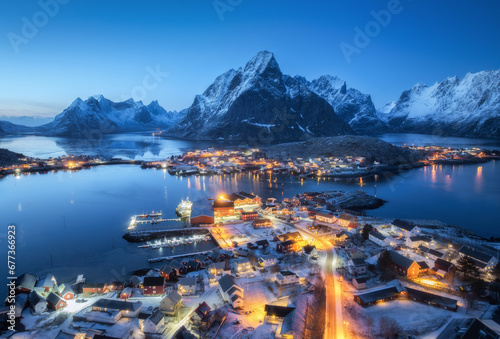 Aerial view of snowy village, islands, rorbu, city lights, blue sea, rocks and mountain at night in winter. Beautiful landscape with town, street illumination. Top view. Reine, Lofoten islands, Norway