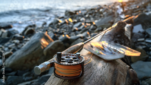 A fly fishing rod and an open fly fishing box lie on the sea rocks at sunset.