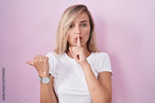 Young blonde woman standing over pink background asking to be quiet with finger on lips pointing with hand to the side. silence and secret concept.