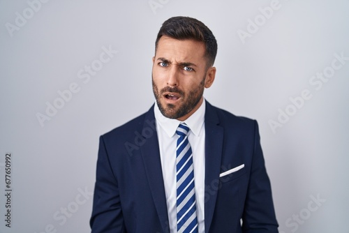 Handsome hispanic man wearing suit and tie in shock face, looking skeptical and sarcastic, surprised with open mouth