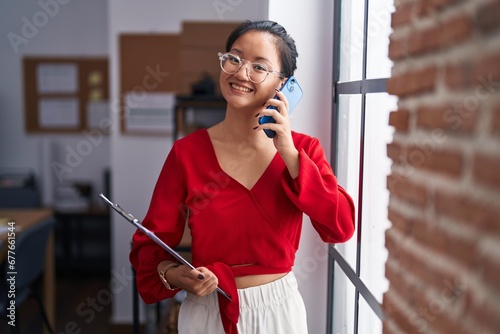 Young chinese woman business worker talking on smartphone holding clipboard at office