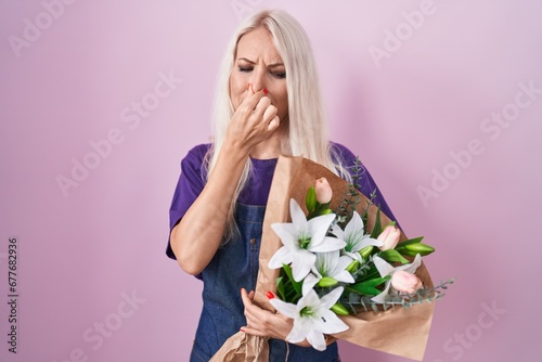 Caucasian woman holding bouquet of white flowers smelling something stinky and disgusting, intolerable smell, holding breath with fingers on nose. bad smell