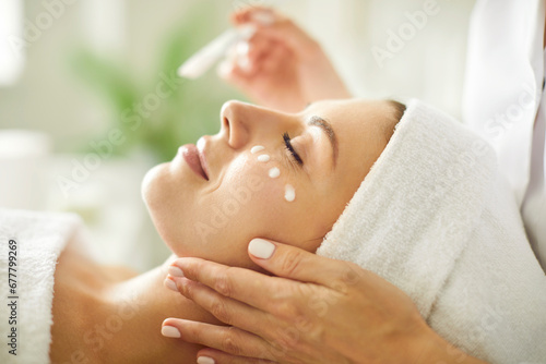 Close up beautician's hands applying anti-aging facial cream on woman client face to prevent wrinkles in spa salon. Skin care, cosmetic procedures for facial care and beauty treatment concept.