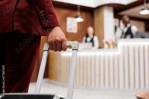 White collar worker with luggage arriving at hotel reception lobby, preparing to see room reservation. Young adult travelling on business meetings, carrying suitcase internationally. Close up.