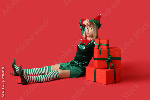 Tired woman in elf costume with Christmas gift boxes on red background