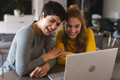 Happy biracial lesbian couple sitting at dining table having laptop video call at home