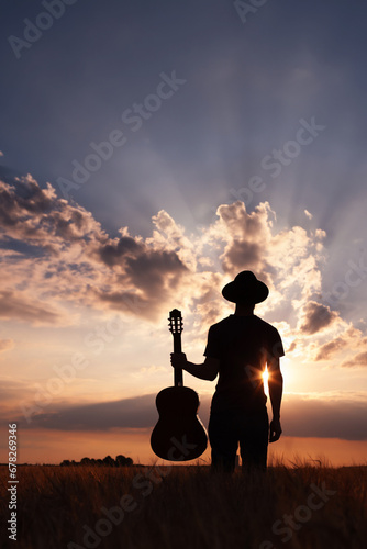 musician standing with guitar at sunset field, music background, silhouette of man artist with guitar and hat