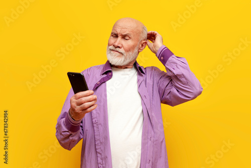 puzzled old bald grandfather in purple shirt uses smartphone on yellow isolated background, confused old pensioner