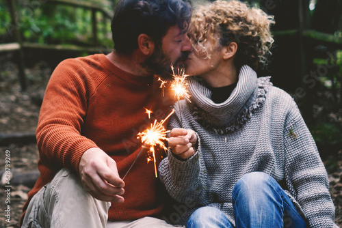 Romantic adult couple kissing in love holding fire sparklers sitting in the nature park enjoy outdoor relationship and leisure activity together. Concept of happy people enjoying vacation