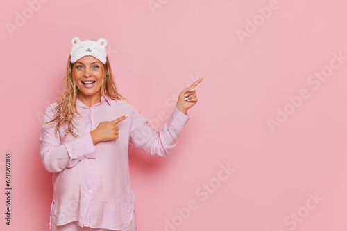 Young smiling pregnant woman dressed in stylish pink pyjamas pointing aside with indes fingers, good mood concept, copy space