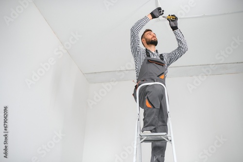 Repairman or professional electrician in workwear installing light spots, standing on the ladder in the white living room